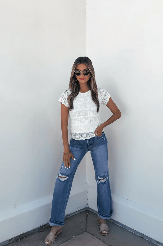 Woman in sunglasses, Amara Cream High Neck Lace Top, ripped blue jeans, and sandals stands against a white wall, hands in pockets.