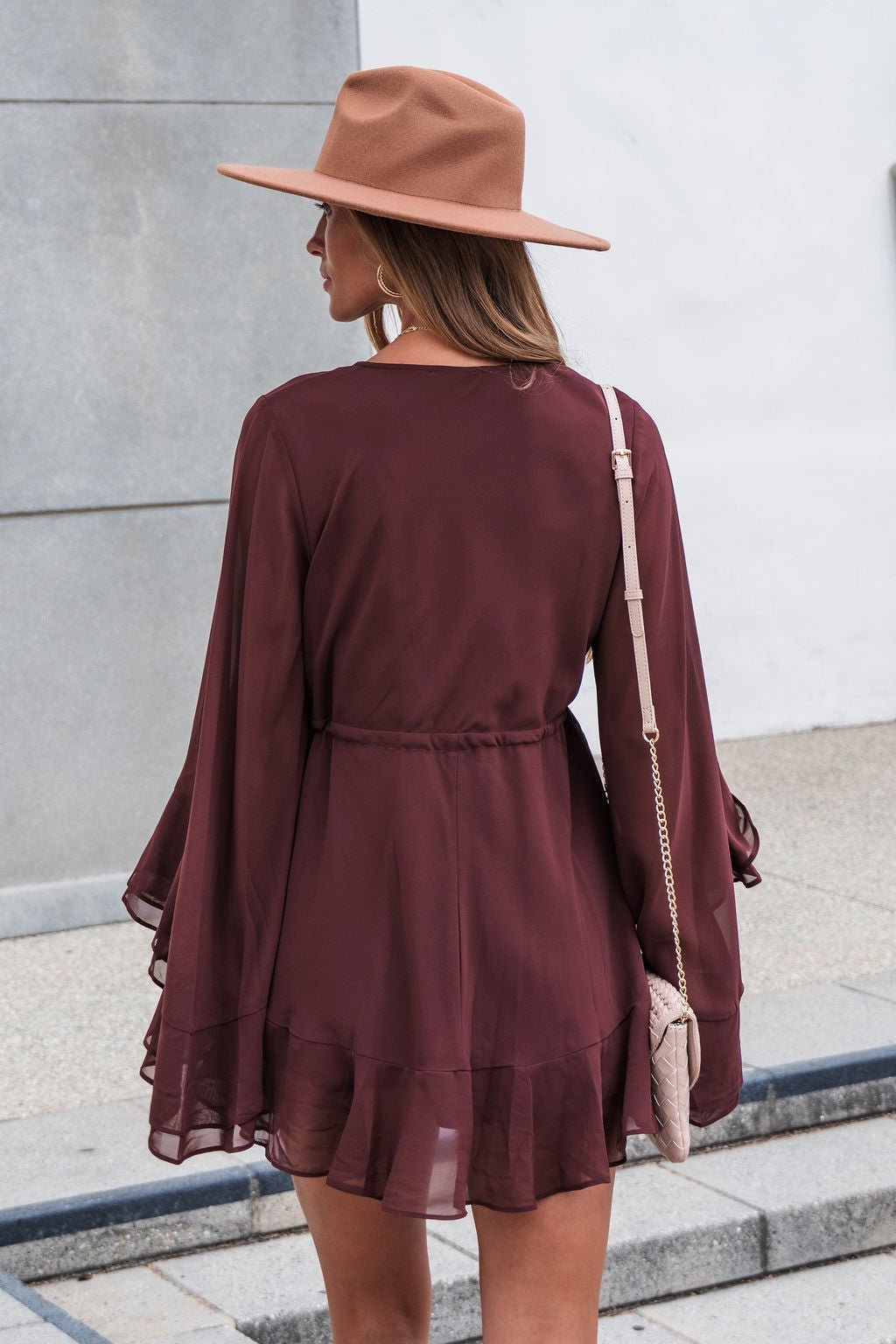 Woman in a brown hat and Amara Ruffled Mini Dress - Brown stands outside, carrying a small beige purse over her shoulder.