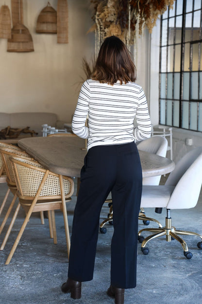 A woman in a striped top and Anabelle Black Pleated Trousers stands by a wooden table in a modern, stylish room.