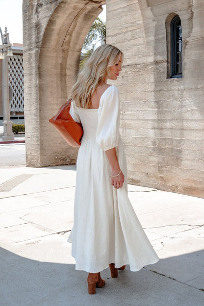 A woman in the Angelica Natural Ruched Midi Dress stands by a stone archway, holding a brown clutch and wearing brown heels.