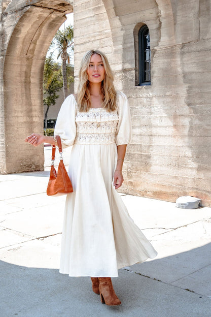 A woman in the Angelica Natural Ruched Midi Dress stands outdoors by stone arches, holding an orange handbag.