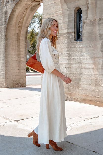 Woman smiles outside by a stone wall, wearing the Angelica Natural Ruched Midi Dress on a sunny day.
