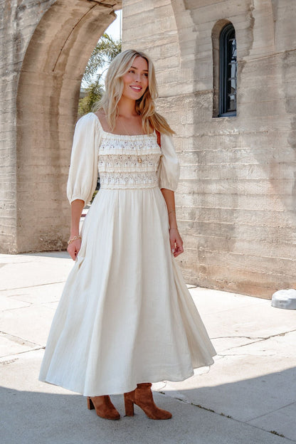 Woman wears the Angelica Natural Ruched Midi Dress, cream cotton with puff sleeves, styled with brown boots outside a stone building.