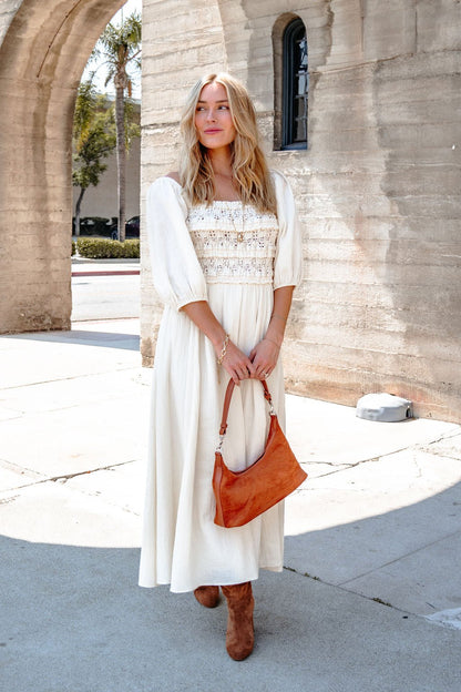 A woman wears the Angelica Natural Ruched Midi Dress and brown boots, holding a brown handbag by a sunlit stone archway.