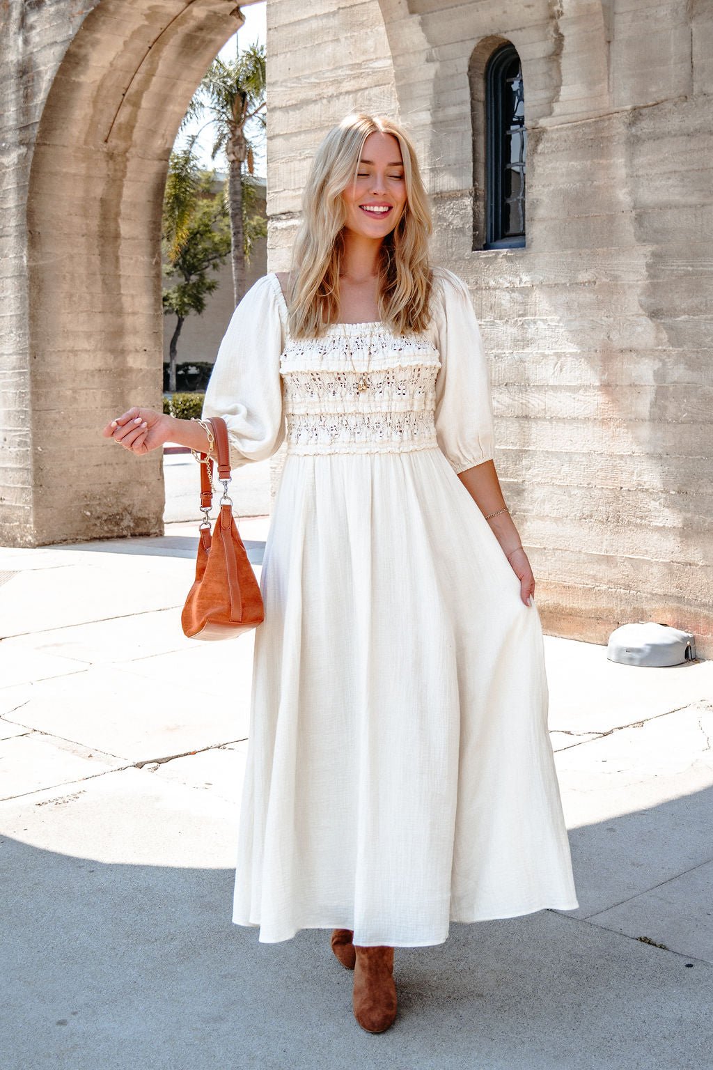 Woman in the Angelica Natural Ruched Midi Dress holds a brown handbag, smiling outside by an arched stone building.