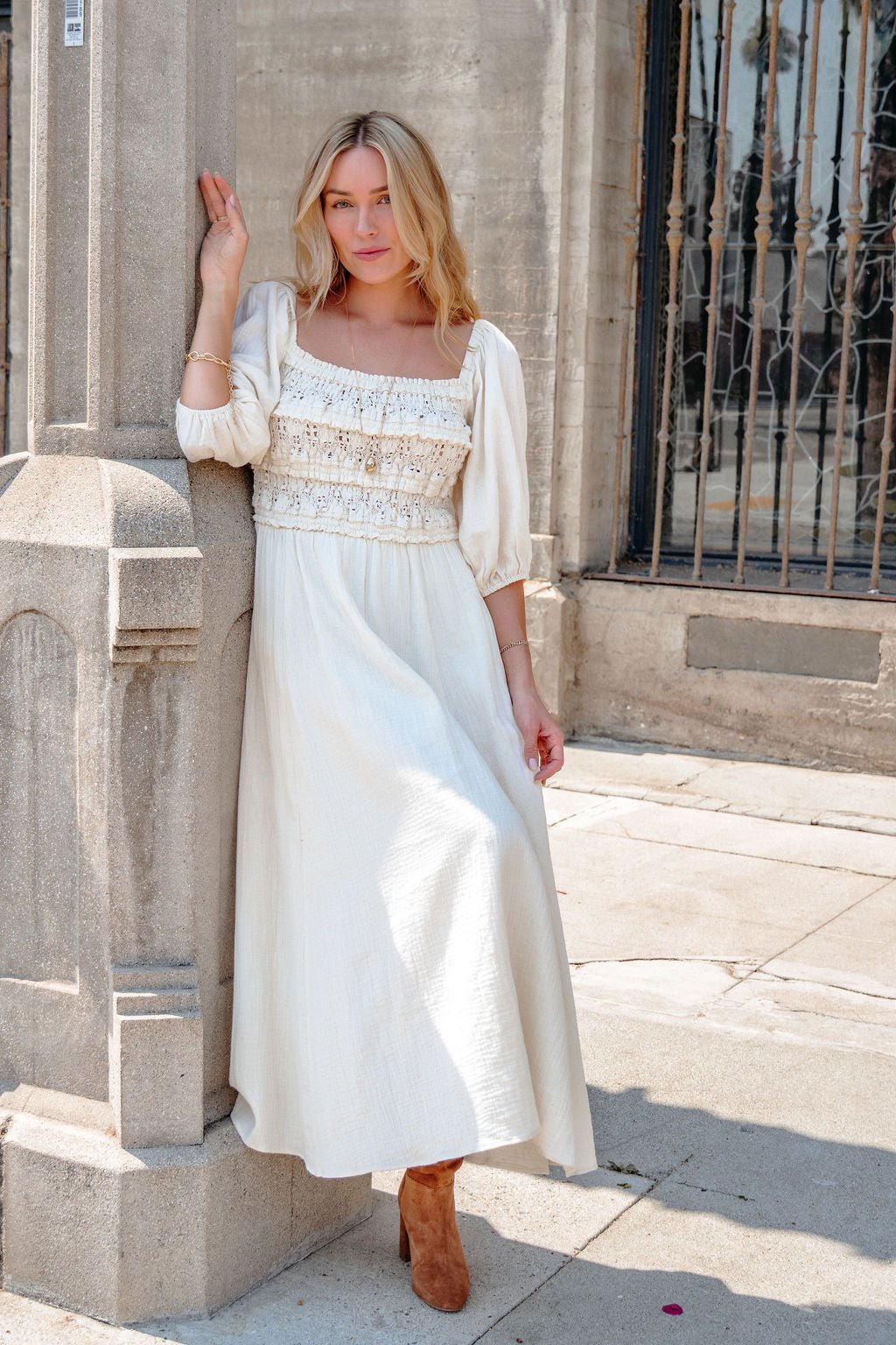 Woman in the Angelica Natural Ruched Midi Dress leans against a stone pillar outside on a sunny day.