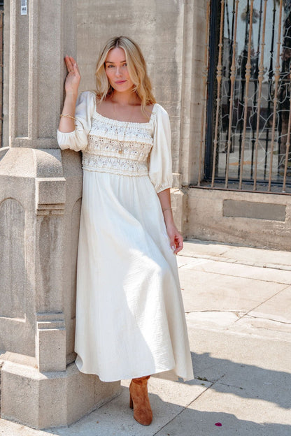 Woman in the Angelica Natural Ruched Midi Dress leans against a stone pillar outside on a sunny day.