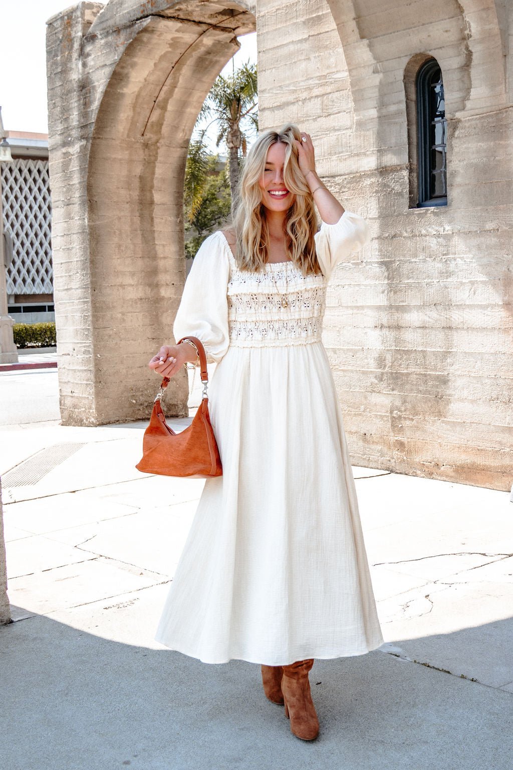 Woman wears the Angelica Natural Ruched Midi Dress, tan boots, and holds a brown purse by stone arches in sunlight.