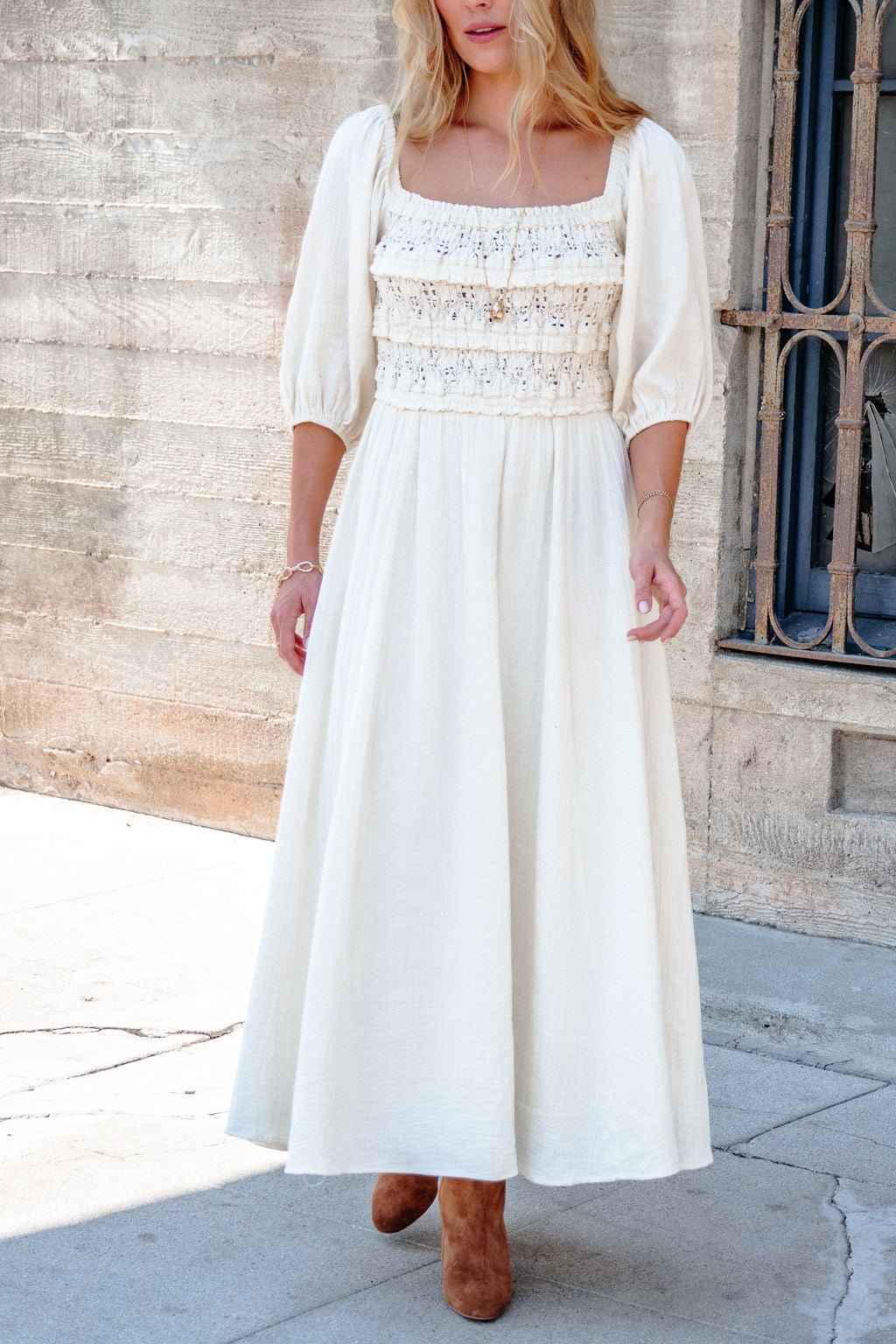 Woman in the Angelica Natural Ruched Midi Dress, standing outdoors by a textured wall, wearing brown boots.