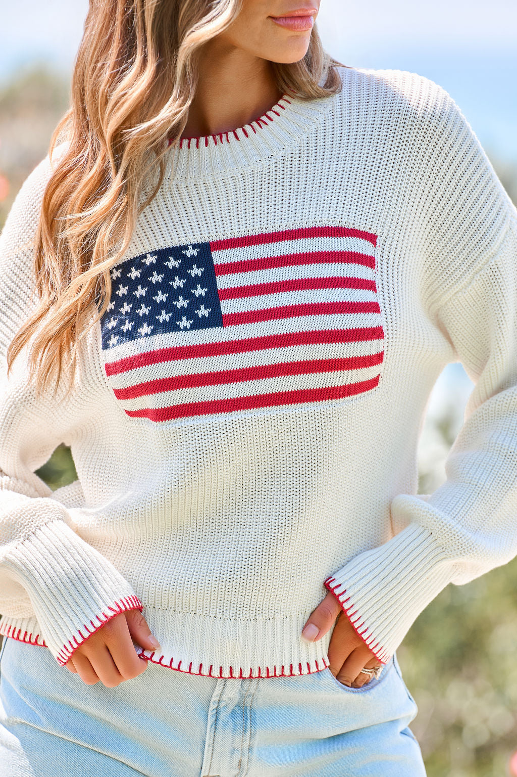 Woman in a relaxed fit Freedom Flag Lightweight Sweater and light blue jeans enjoys the sunlight outdoors.