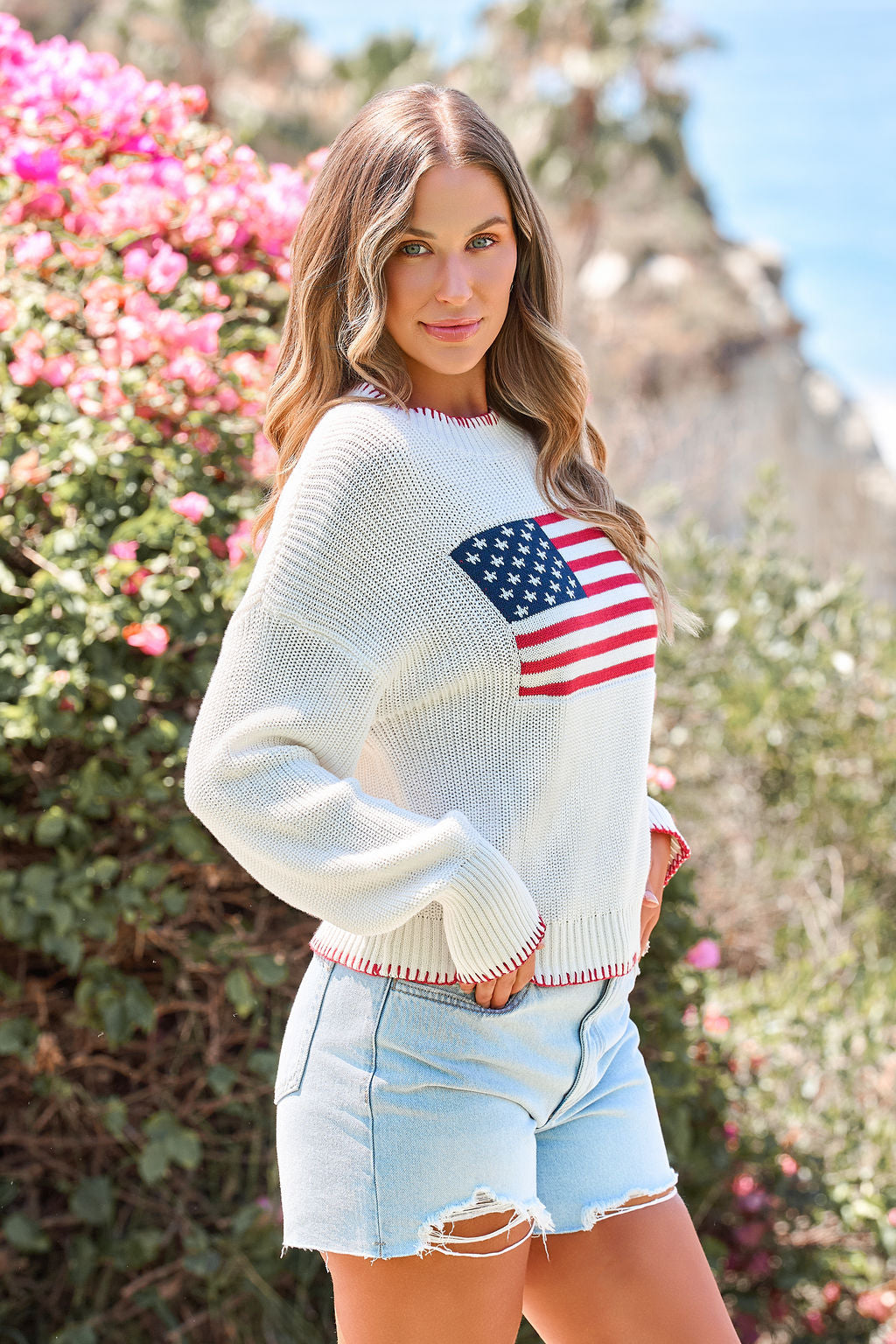 Woman outdoors in a relaxed fit Freedom Flag Lightweight Sweater and light denim shorts, standing by blooming flowers.