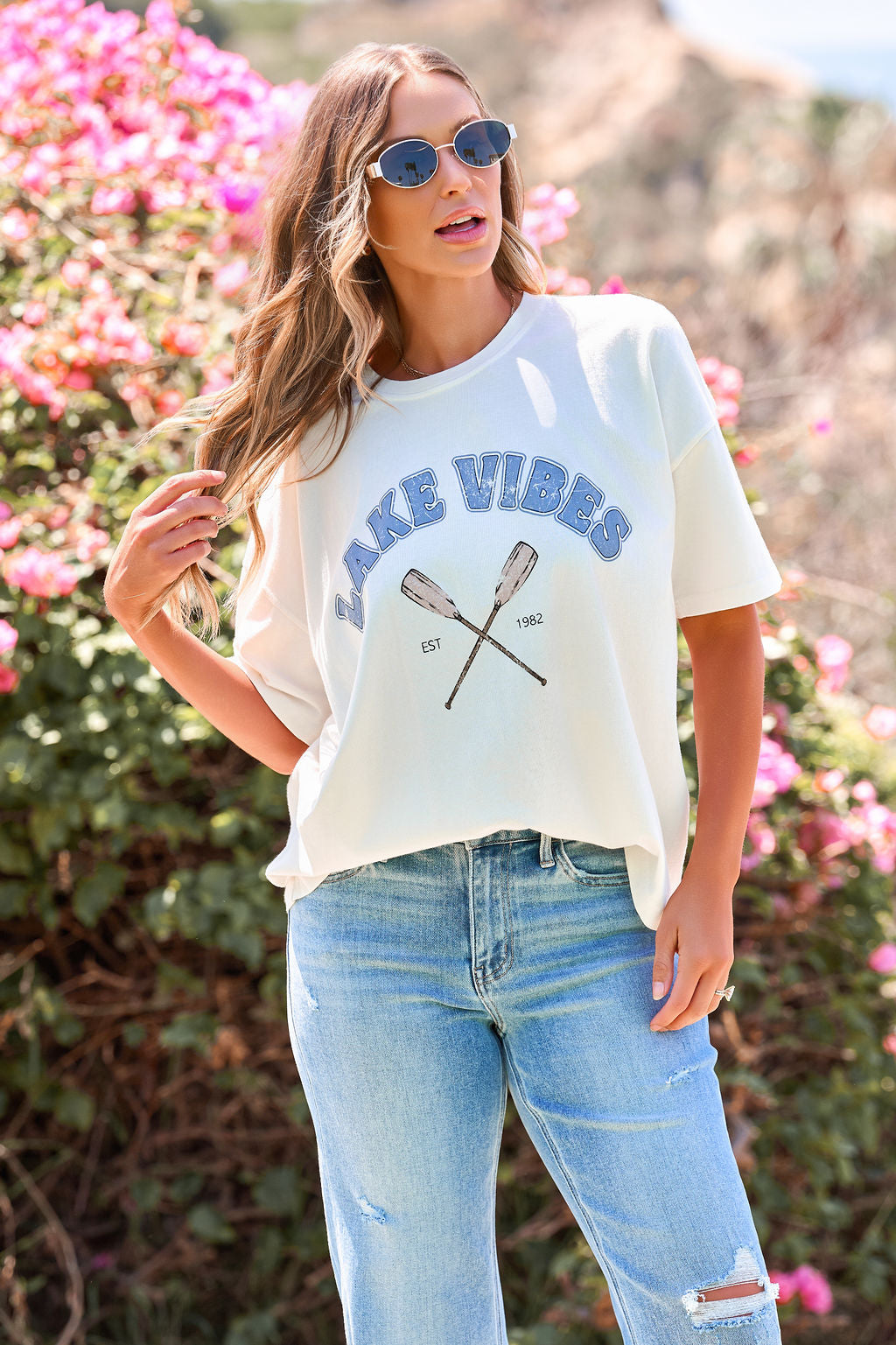 A woman in sunglasses, jeans, and the Lake Vibes Graphic Tee - Ivory stands outdoors near pink flowers on a sunny day.