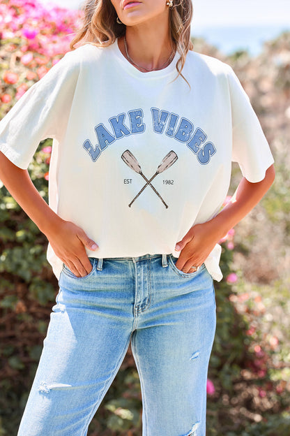 Woman in a Lake Vibes Graphic Tee - Ivory and light blue jeans stands outdoors among flowers and greenery.