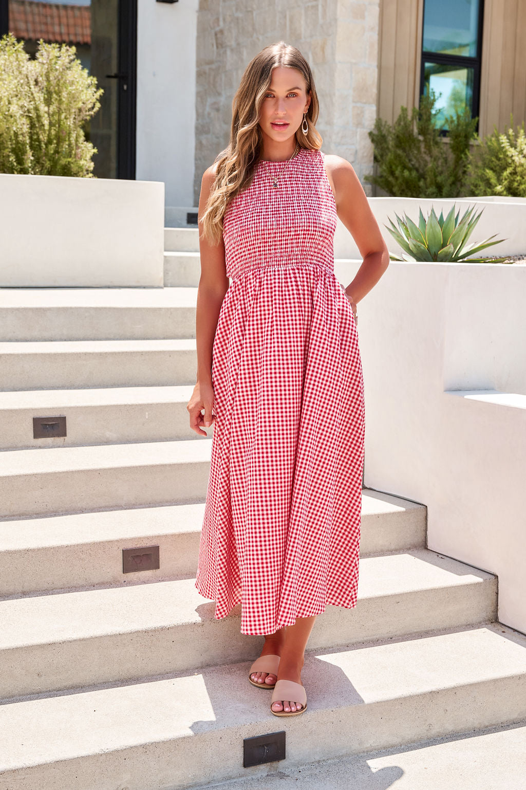 Woman wearing the Gingham Open Back Maxi Dress - Red, standing on outdoor steps by a modern building.