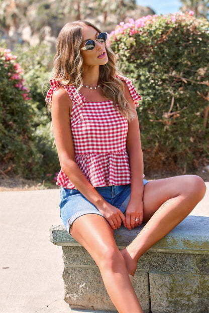 Woman in sunglasses wears the red Gingham Babydoll Tank Top with tie straps and denim shorts, sitting on a stone bench.