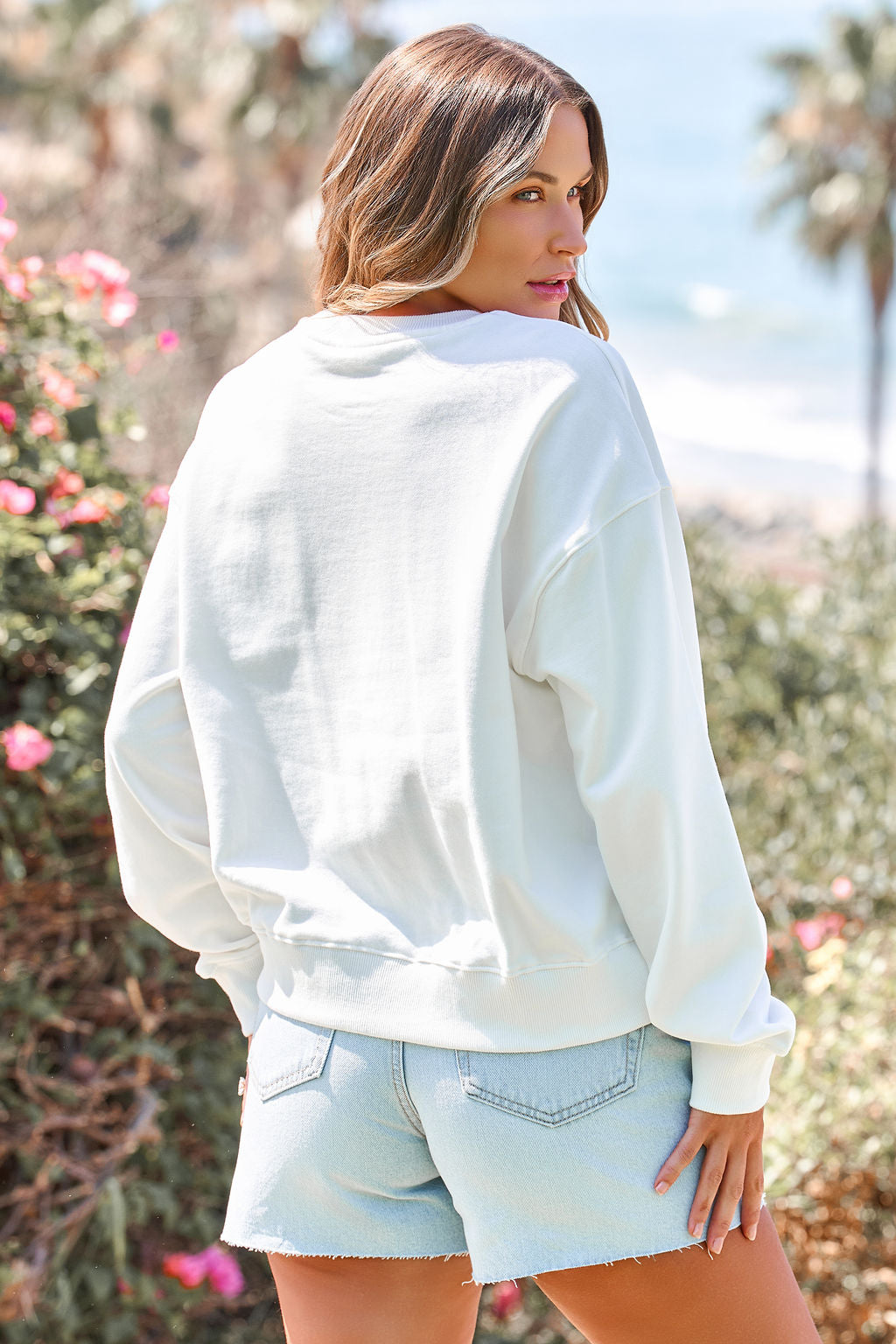 Woman with wavy hair wears the Bold & Free Patchwork Sweatshirt - White and denim shorts, standing outdoors and glancing back.