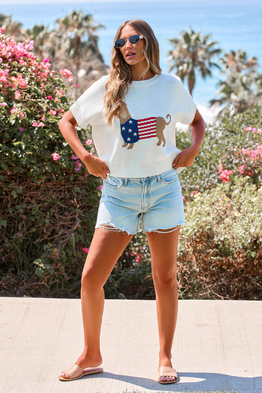 Woman in sunglasses wears the Patriotic Pup Knit Sweater - Cream with denim shorts and sandals by the ocean.