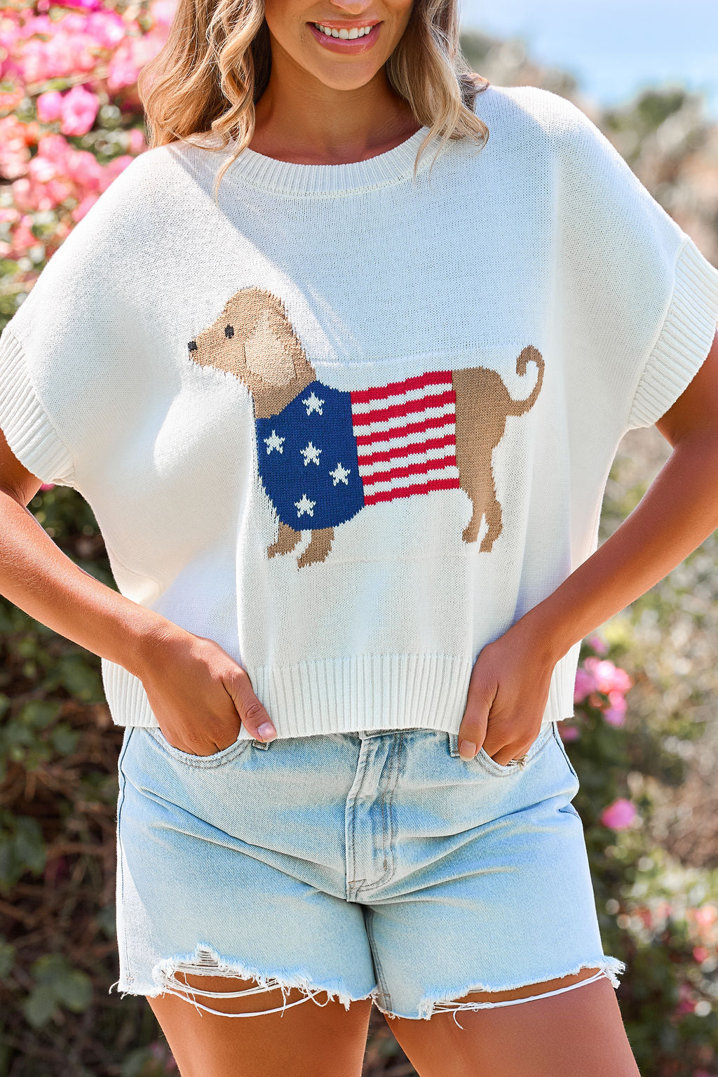 Woman in denim shorts wears the Patriotic Pup Knit Sweater - Cream, featuring a dog and flag, standing outside by flowers.