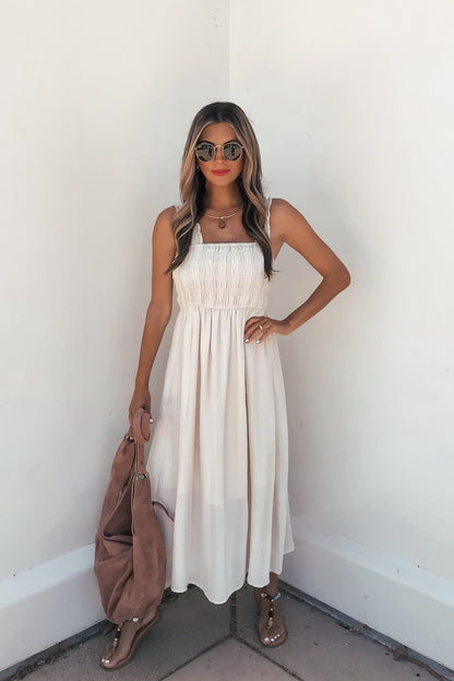 Woman in the Arwen Ruffle Smocked Midi Dress - Natural, sunglasses, sandals, and a brown bag by a white wall.