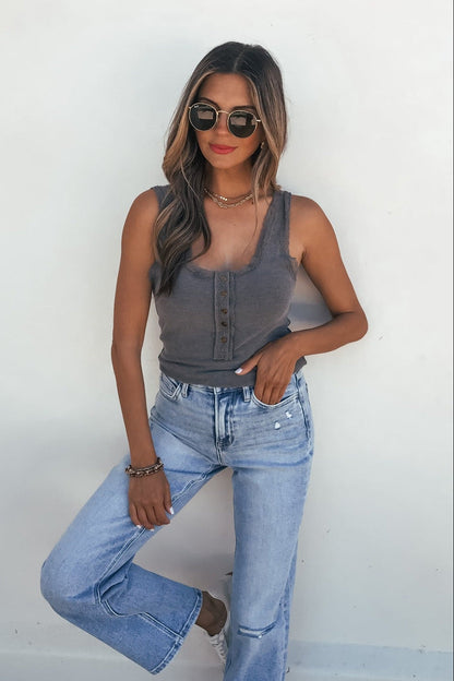 Woman in sunglasses wears the Ashlyn Lace Trim Button Tank – Grey with light blue jeans, posing by a white wall.