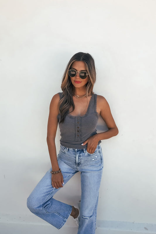 Woman in sunglasses wears the Ashlyn Lace Trim Button Tank – Grey with light blue jeans, posing by a white wall.