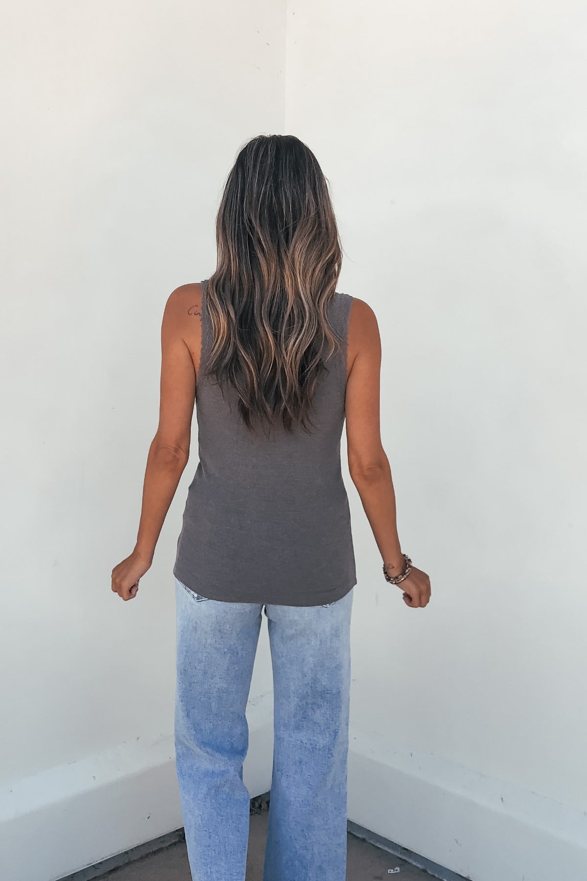 A woman wears the Ashlyn Lace Trim Button Tank in grey and light blue jeans, standing with her long wavy hair facing a white wall.