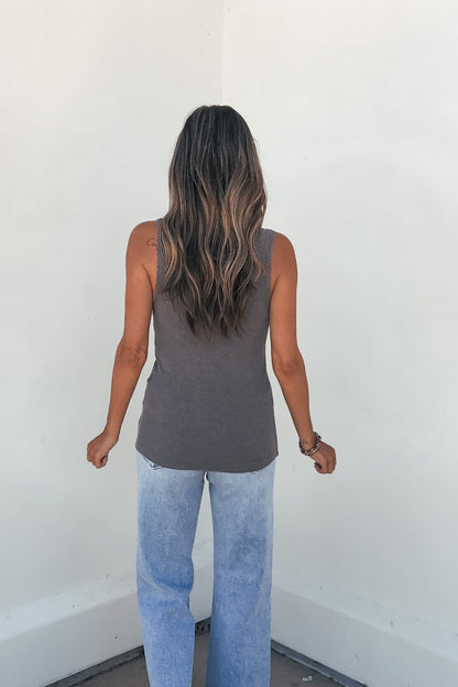 A woman wears the Ashlyn Lace Trim Button Tank in grey and light blue jeans, standing with her long wavy hair facing a white wall.