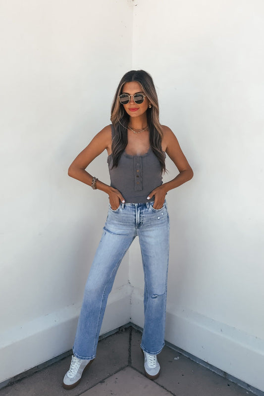 Woman wears sunglasses, Ashlyn Lace Trim Button Tank – Grey, light blue jeans, and white sneakers against a white wall.