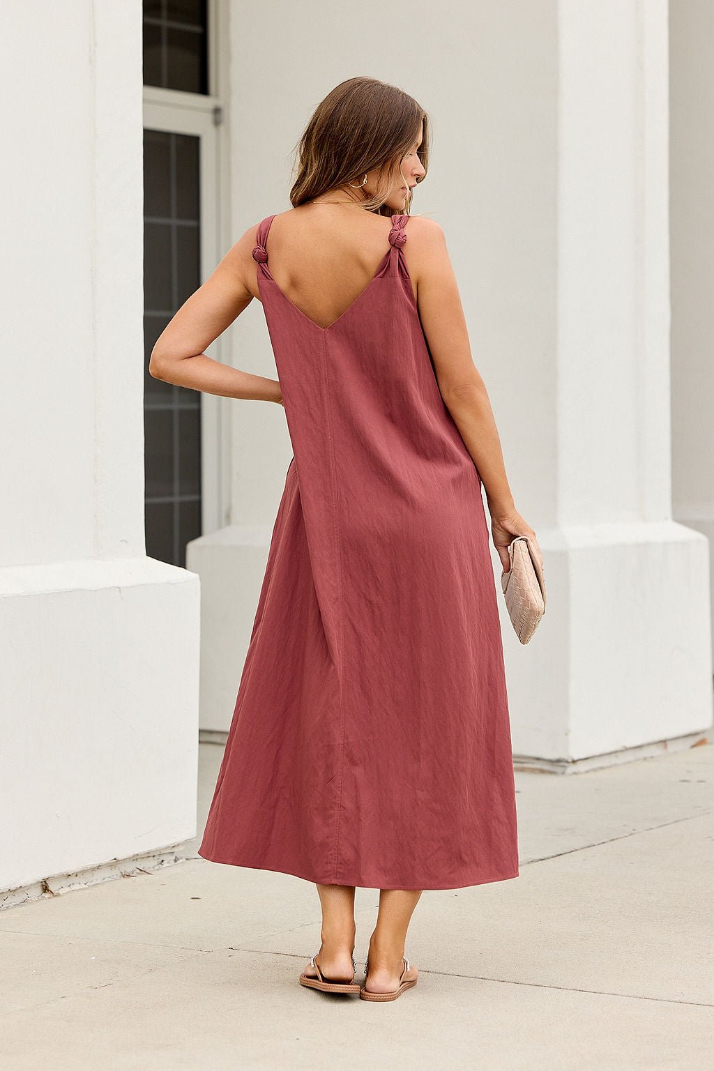 Woman in the Audrey Knot Strap Midi Dress - Burgundy, holding a clutch by a white building outdoors.