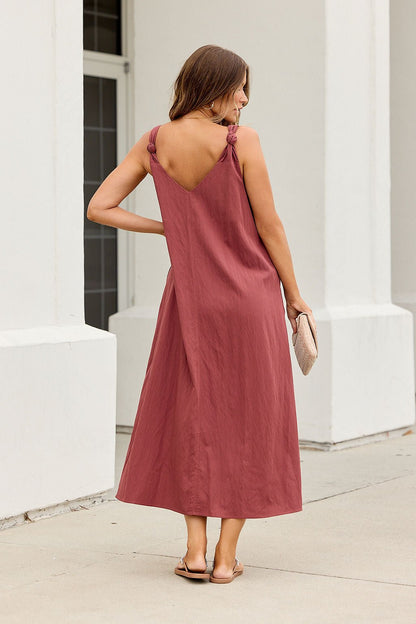Woman in the Audrey Knot Strap Midi Dress - Burgundy, holding a clutch by a white building outdoors.