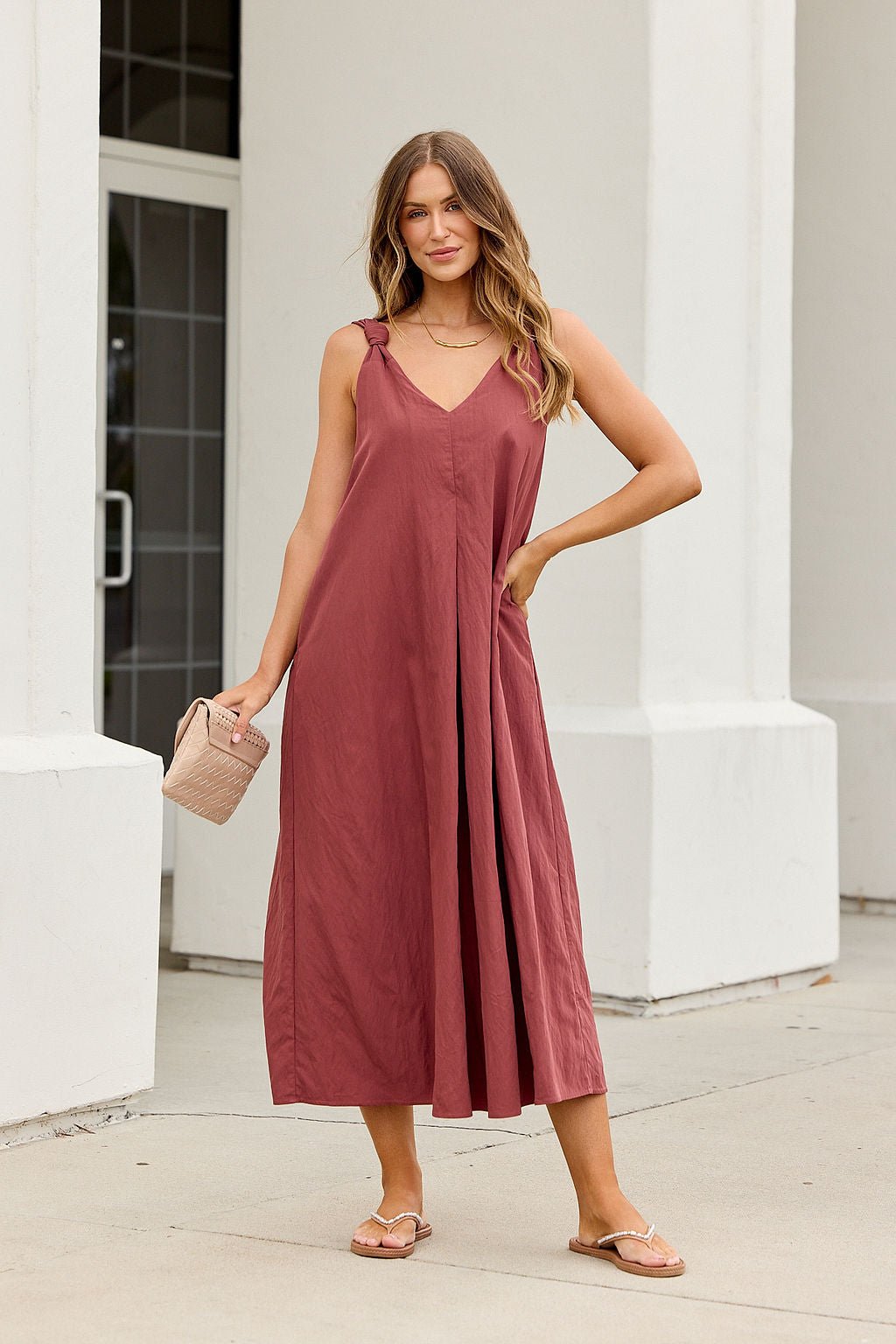 Woman in the Audrey Knot Strap Midi Dress - Burgundy, sandals, and woven bag stands outside a white building.