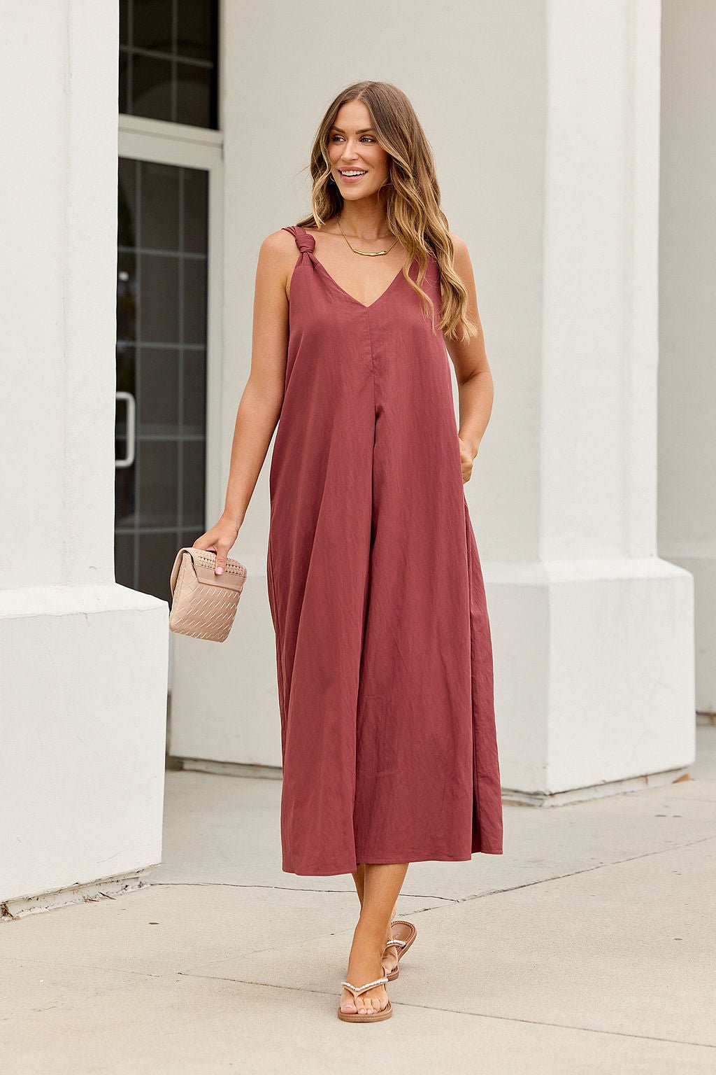 Woman in burgundy Audrey Knot Strap Midi Dress, sandals, wavy hair, and small handbag stands by a white building on the sidewalk.