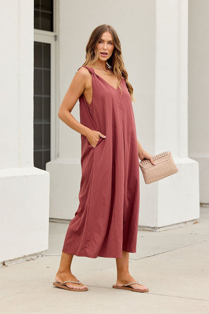 Woman in the Audrey Knot Strap Midi Dress - Burgundy, with pockets, holding a beige clutch, wearing sandals on a city sidewalk.