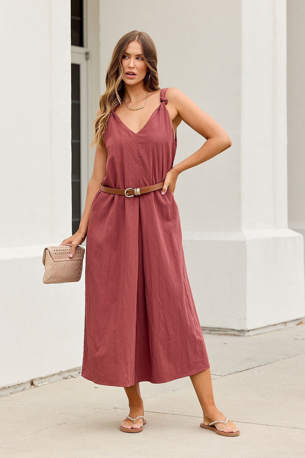 Woman in the Audrey Knot Strap Midi Dress - Burgundy holds a beige clutch while standing by a white building.