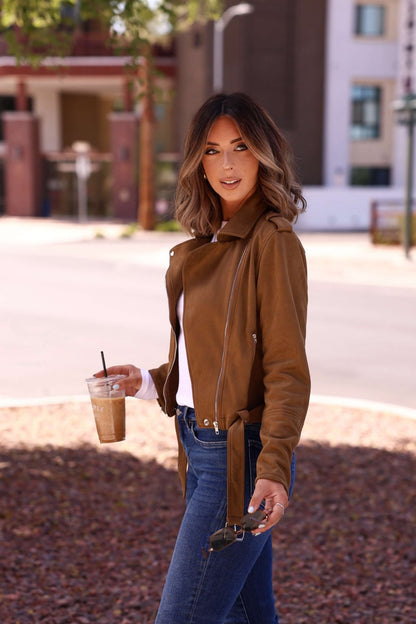 Woman outdoors in the Audrey Taupe Vegan Suede Moto Jacket and jeans, holding iced coffee and sunglasses, looking at the camera.