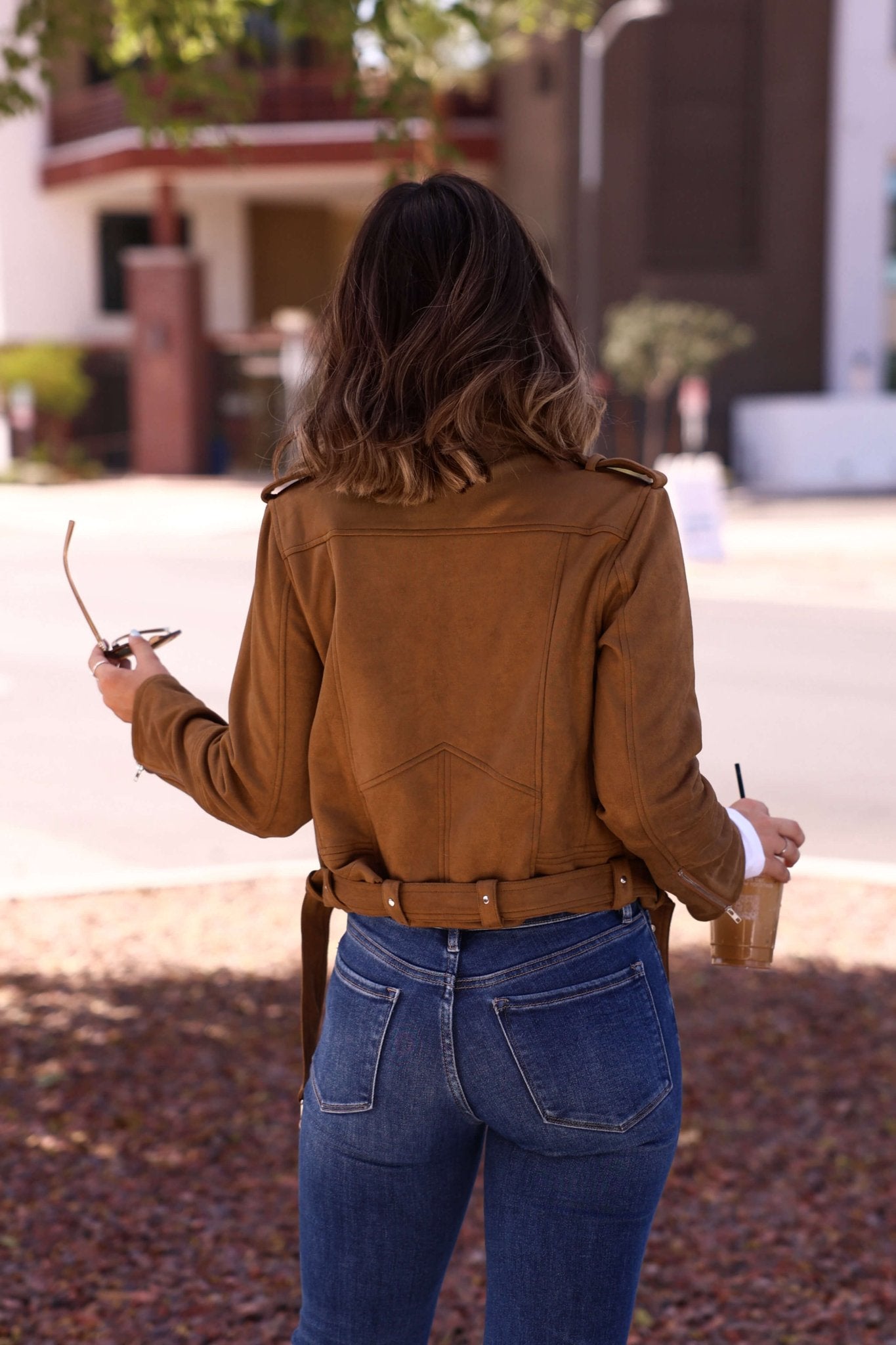 Woman with wavy hair stands outdoors, facing away, in the Audrey Taupe Vegan Suede Moto Jacket and jeans, holding sunglasses and a drink.