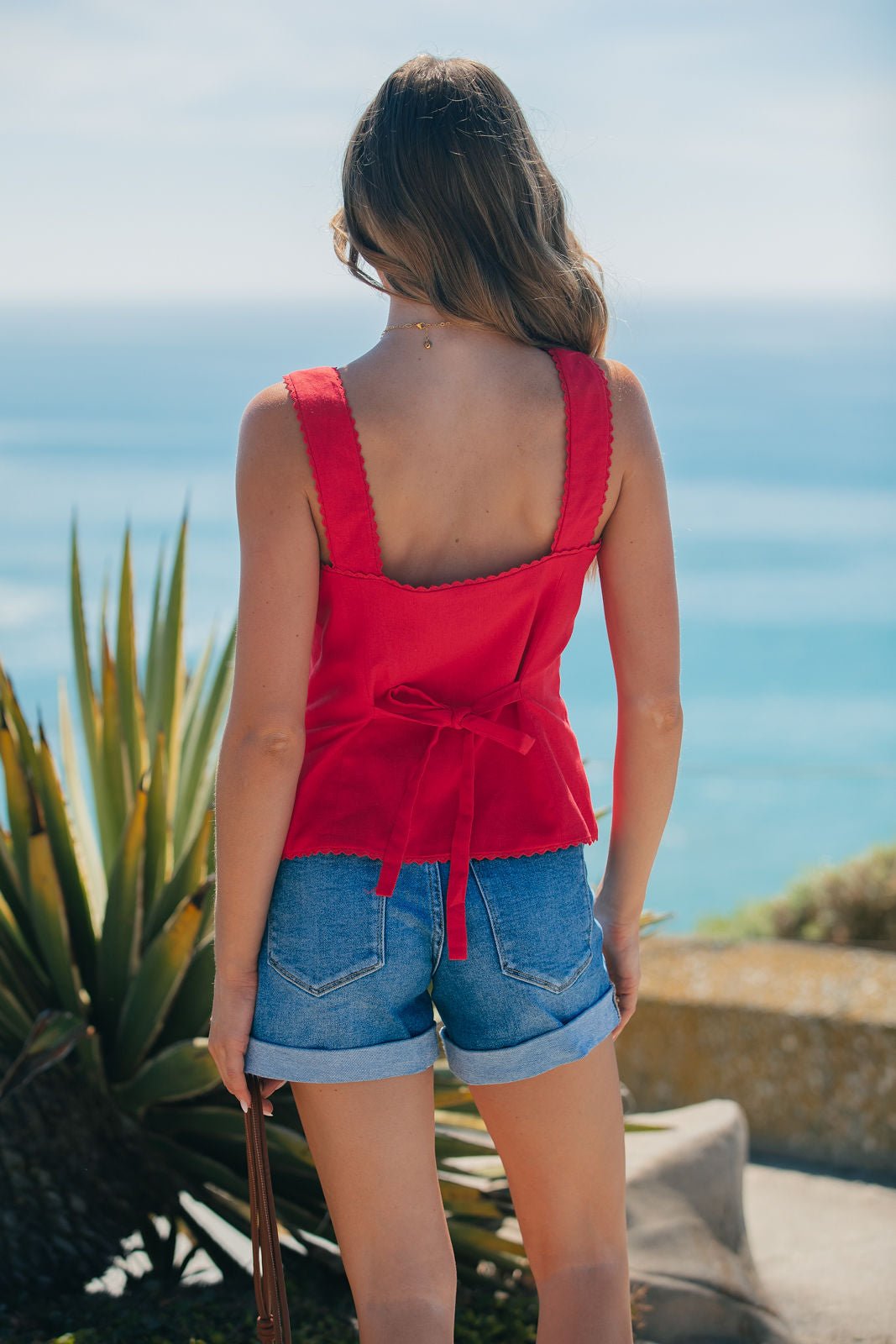 A woman in an Aureum Lace Trim Linen Tank Top - Red and denim shorts stands outdoors, facing the ocean with nearby plants.