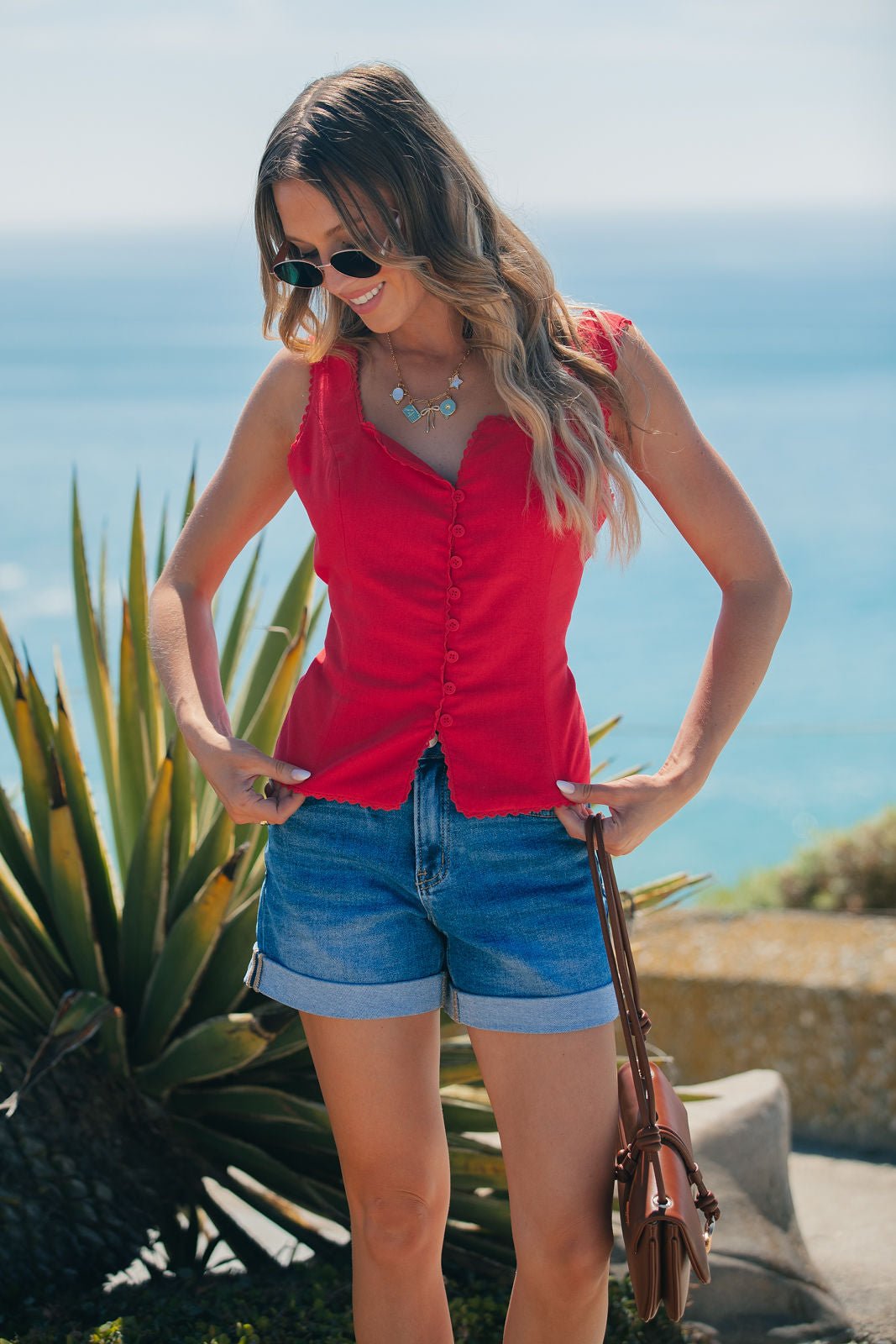 Woman in a red Aureum Lace Trim Linen Tank Top and denim shorts stands by the ocean, wearing sunglasses and holding a brown purse.