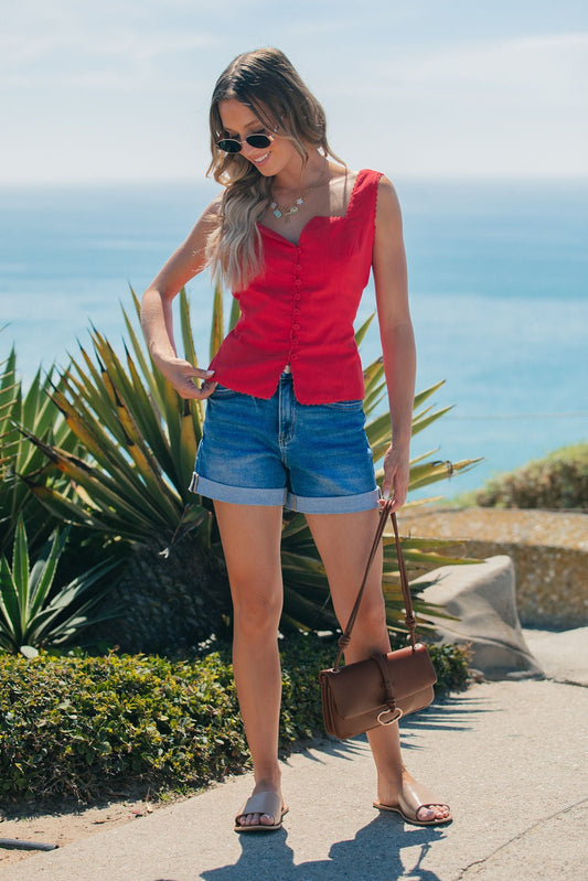 Woman in the Aureum Lace Trim Linen Tank Top - Red, denim shorts, and sandals stands near the ocean with a brown purse.