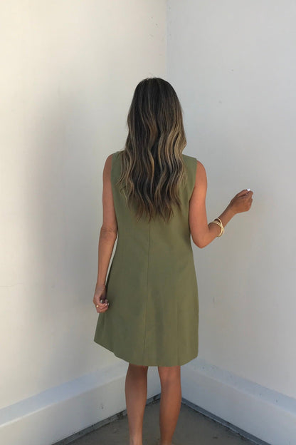 A woman with long wavy hair models the Aureum Uptown Olive Grove Mini Dress, standing with her back to a plain wall.