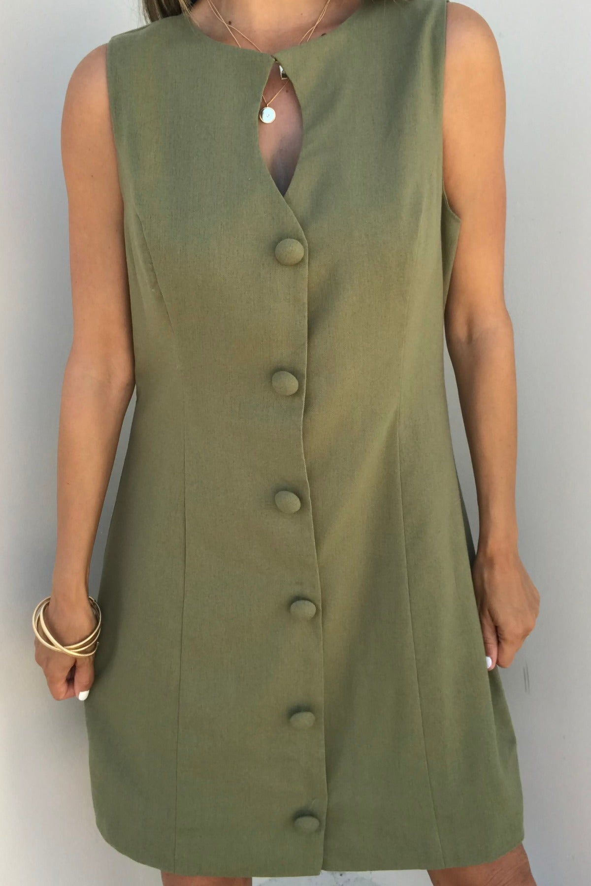 Woman wears the Aureum Uptown Olive Grove Mini Dress—sleeveless, olive green, front buttons, keyhole neckline, and gold bangles.