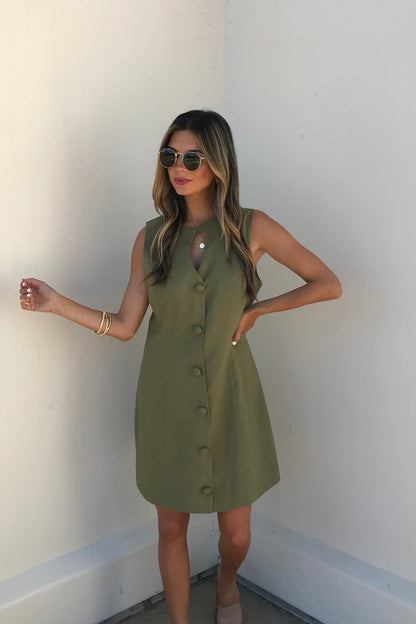 Woman in sunglasses and the Aureum Uptown Olive Grove Mini Dress stands against a white wall.