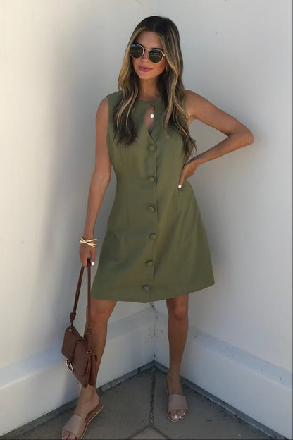 Woman in the Aureum Uptown Olive Grove Mini Dress, sunglasses, nude sandals, brown purse, posing by a white wall.