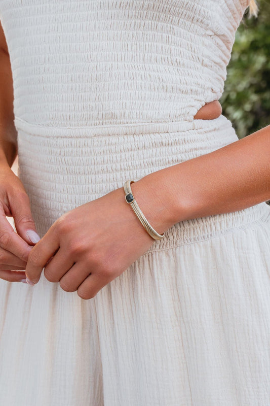 A person in a textured white dress wears the Aurielle Smoky Stone Bracelet—a modern gold piece with a dark stone accent.