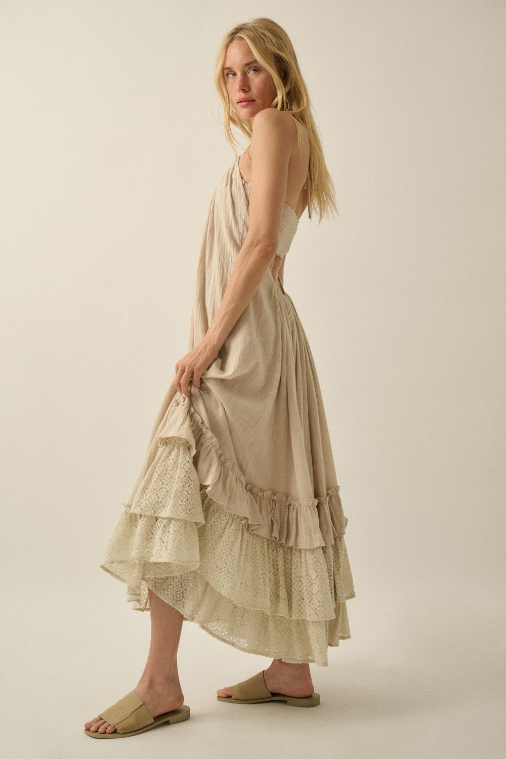 Woman in the Aurora Natural Mixed Lace Maxi Dress with ruffles and halter neckline stands sideways, wearing tan slide sandals.