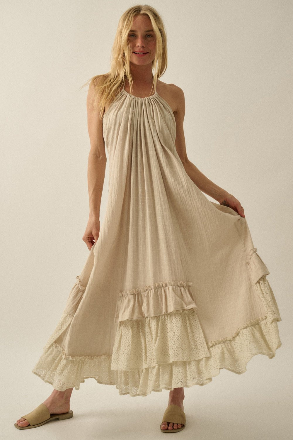 Woman in the Aurora Natural Mixed Lace Maxi Dress with a ruffled hem and halter neckline, smiling as she holds out the dress sides.