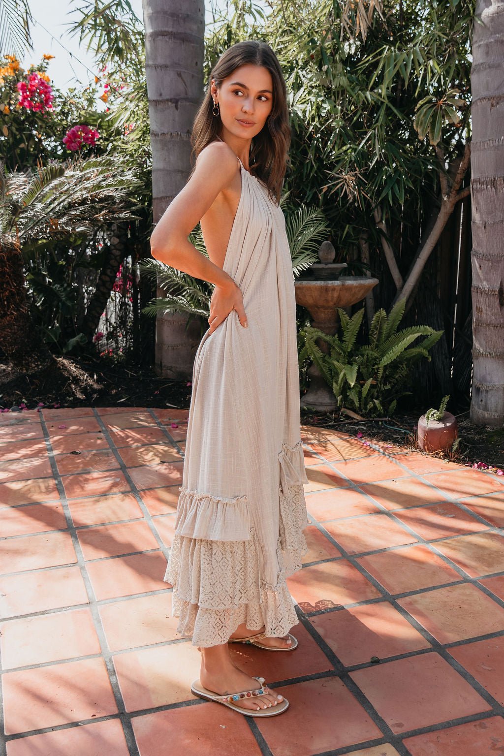 Woman in the Aurora Natural Mixed Lace Maxi Dress stands on a tiled patio, surrounded by lush greenery and tropical plants.