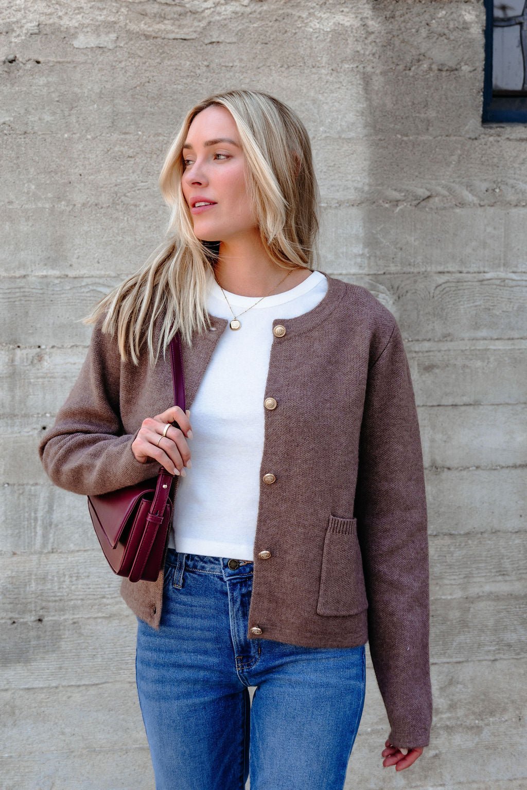 A woman in jeans, a white shirt, and the Autumn Button Front Sweater Cardigan in brown stands by a concrete wall with a maroon purse.