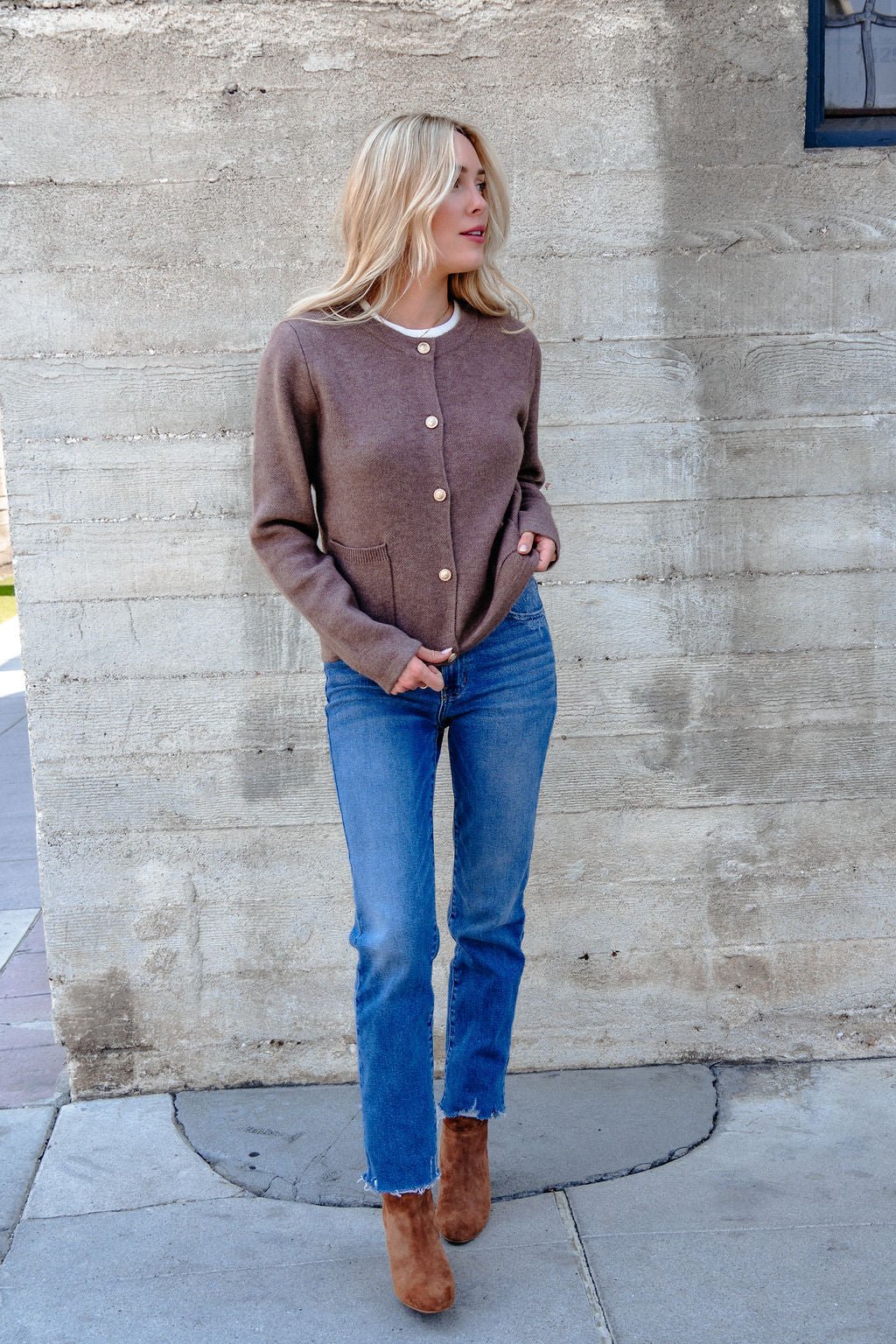 Woman in the Autumn Button Front Sweater Cardigan - Brown, blue jeans, and boots stands by a concrete wall, looking left.