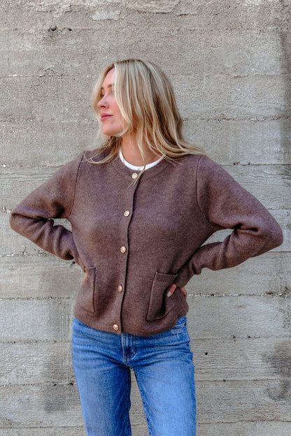 Woman wears the Autumn Button Front Sweater Cardigan in brown with blue jeans, standing by a concrete wall, looking to the side.